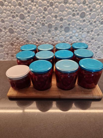11 standard jars with blue lids & 1 smaller jar with white lid full of red currant jam, resting on a bamboo cutting board in front of a random white grey silver circle backsplash