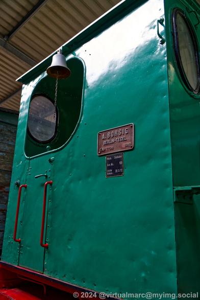 A photo of the drivers cab of a green steam locomotide from the side outside. There is a bell near the window. A sign near the window reads:
A. Borgsig
Berlin-Tegel
F.No 7794  1910

Werk: Südkabel
Betr. Nr.: 120
Letzte Untersuchung: 22.3.72