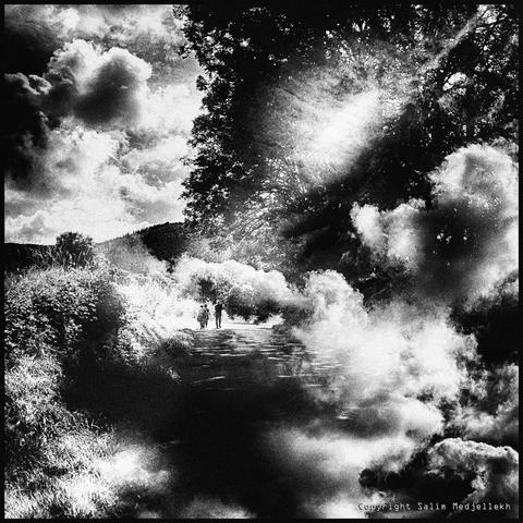 Photographie en noir et blanc de trois personnes marchent loin sur un chemin entourés de nuages qui les enveloppent avec le soleil qui les transperce.
Black and white photograph of three people walking far along a path surrounded by clouds that envelop them with the sun shining through.