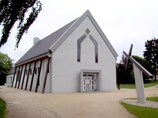 The image shows a modern architectural building, likely designed for a communal or religious purpose, given the presence of a cross structure nearby. The building has a distinctive geometric design with a slanted roof and a facade that includes angular elements. There are also poles or beams supporting the structure. The surrounding area appears to be landscaped with gravel and grass.

Image Credits: Wikimedia / Methusalem in der Wikipedia auf Deutsch / Public domain