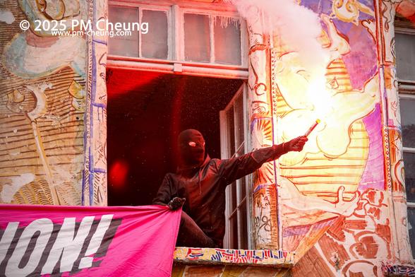 Von einem Fenster des Autonomen Zentrums sitzt eine vermummte Person, der eine bengalische Fackel gezündet hat. Es handelt sich hierbei um eine Solidaritätsbekundung mit den Demonstrant*innen, die so eben eingetroffen sind.