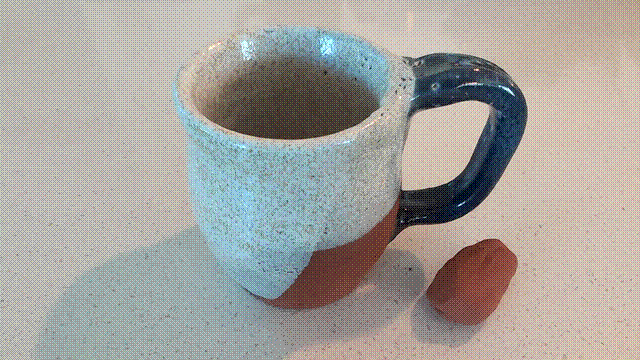 A mug with speckled white glaze  over most of it. The handle is a dark mottled blue. The bottom corner shows bare, red clay. A small lump of unglazed red clay sits next to it.
