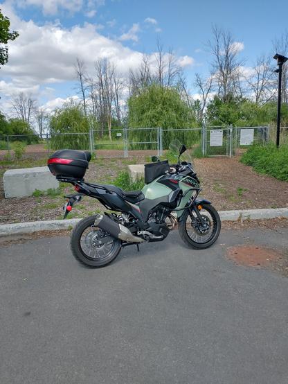 Image shows a kaki coloured Kawasaki Versys X 300 adventure motorcycle on a street with a chain link fence behind it and beyond that some trees and a blue sky with some puffy clouds.