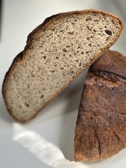A close-up view of a sliced loaf of dark, artisanal bread, showcasing its texture and air pockets, with a rustic crust and a section of the whole loaf visible in the background.