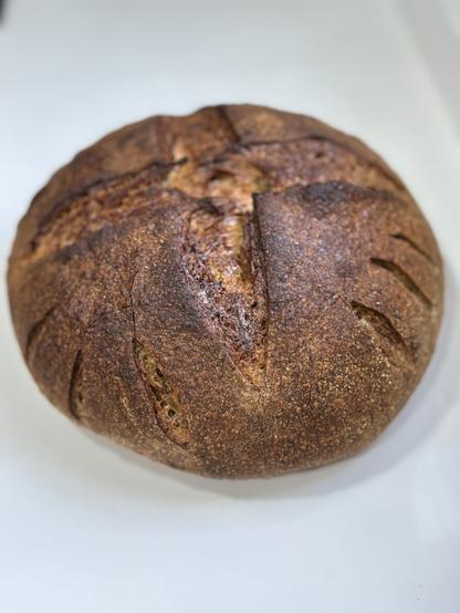 A round loaf of crusty bread with a dark, textured crust and scored design on top, placed on a white surface.