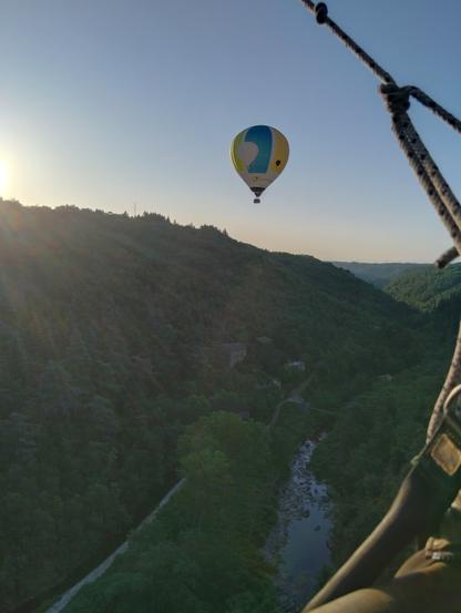 Vue de la nacelle sur une autre montgolfière qui survole une vallée verte de végétation avec vous d'eau en contrebas. Le soleil levant sur la gauche crée une surexposition en haut de la colline.