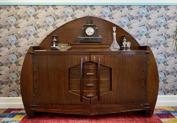 Buffet bas en bois sombre en forme d’ogive, d’un style qui était moderne dans les années 10 ou 20. Au centre, la partie à la jonction des deux portes forme un hexagone coupé par deux grandes poignées verticales. Plusieurs objets (carafe, verres, pendulette) sont posées sur le dessus du meuble.