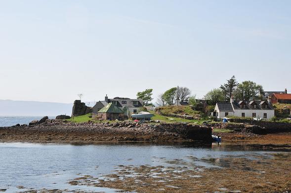 The harbour at Camusterrach on the Applecross peninsula. The image shows a view over water with the tide well out to part of a village on a promontory with the walls of an old harbour and boats moored and on land. The sky is light blue.