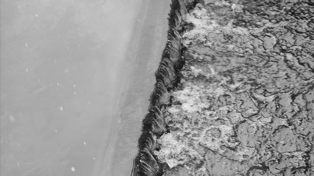 Water flowing over a sharp-edged weir, creating ripples and white foam on the right side of the image. Smooth, calm water lies to the left of the weir in a contrasting texture. The image is in grayscale, highlighting the contrast between the turbulent and still water.