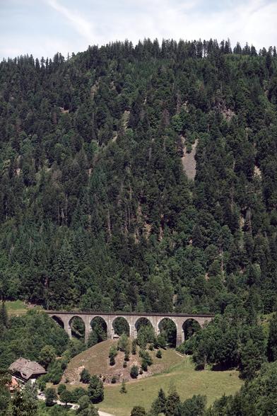 Im unteren Bild-Teil ist ein altes Bahn-Viadukt mit 7 Bögen zu sehen, auf dem ein Bahngleis gebaut ist. Dahinter baut sich ein steilerer Berghang mit Wald auf.