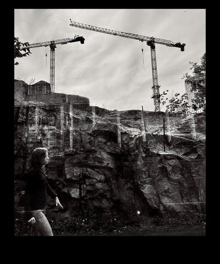 A person walks past a rocky landscape with two construction cranes towering in the background under a cloudy sky, captured in black and white.