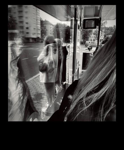 A monochrome image of a bus stop, featuring hair and a face in an advertisement, also reflections of persons and nearby urban landscape can be seen.