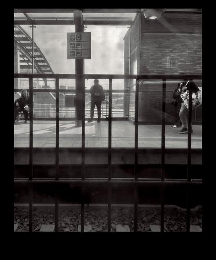 A black-and-white image of a metro station platform with people waiting, framed by a metal railing.