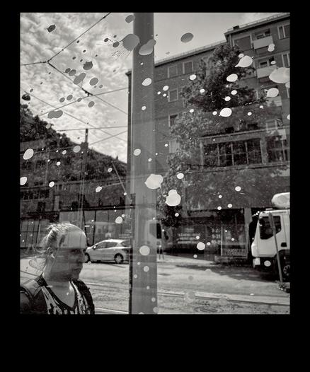 An urban scene viewed through a glass of a bus stop, featuring a passerby, scattered white paint splatters and a glimpse of a street with parked cars.