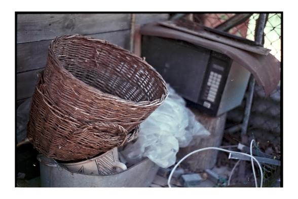 A worn wicker basket sits atop a metal container in a cluttered outdoor corner, next to an old microwave and scattered household items. The colors are muted and slightly faded, captured on expired color film using a Minolta Dynax 7