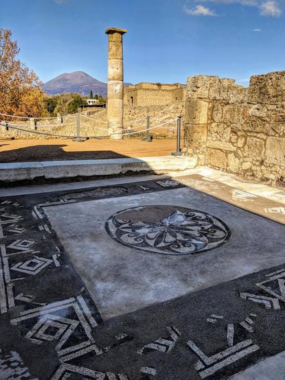 A sunlit, wide-angle shot of Roman ruins in Pompeii, Italy. In the foreground, an ornate black-and-white mosaic floor with geometric patterns and a central circular motif is visible. Beyond it, a lone, fluted stone column stands tall amidst crumbling stone walls and foundations. In the distance, Mount Vesuvius rises under a clear blue sky with sparse clouds.