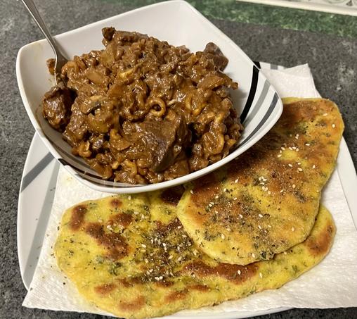 a plate on a dark countertop. on the plate is two pieces of pan-fried flatbread laying on kitchen paper. the flatbread is crusted with herbs and spices. on top is a bowl of rich brown stew, with a fork poking out of it.