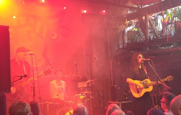 die Band "Acht Eimer Hühnerherzen": echts Johnny Bottrop mit Akustik Bass, rechts die Sängerin Apocalypse Vega mit Akkustkgitarre. in der Mitte der Schlagzeuger Benedikt Diktator. im Vordergrund ein rotes Banner mit Herz, das Band Logo, im Hintergrund das Logo vom Schokoladen. das Licht ist rötlich orange