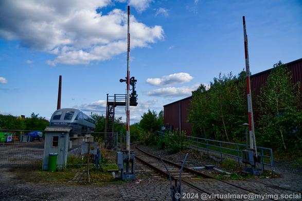 A photo of the railway crossing. The train car hall 2 can be seen on the right. A form signal on a small bridge can be seen behind the railway crossing. Left of it you can see the Transrapid 07 from t he front.