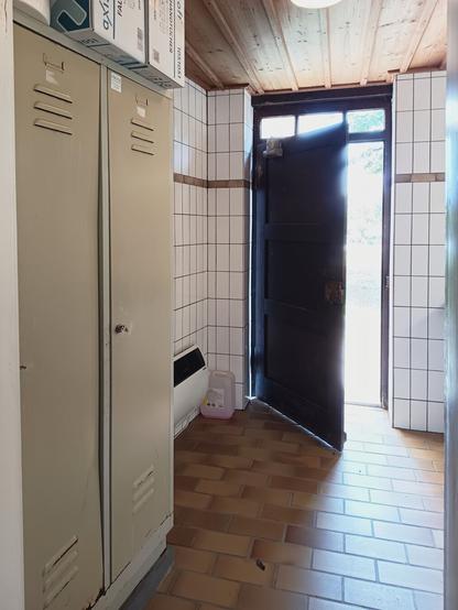 A narrow, tiled room with a wooden ceiling and yellow lockers on the left. An open, black door on the right lets in bright daylight, creating a utilitarian and transitional atmosphere.