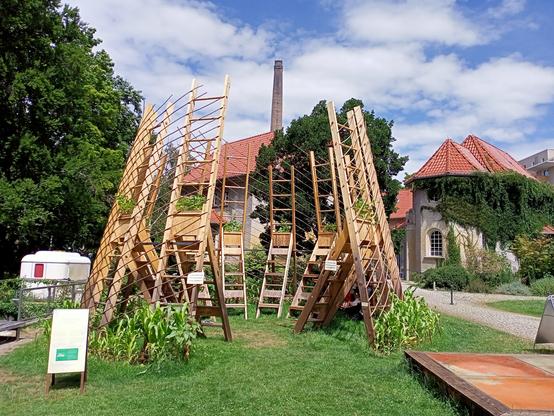 Eine konische Holzkonstruktion, teils bewachsen, innen mit leiterartigen Sitzen versehen, die durch schraubenförmig sich in die Höhe drehende Weidenruten verbunden sind. Das Ganze sieht offen, zart und luftig aus. Im Hintergrund sind Bäume und kleinteilige Backsteinbauten zu sehen.