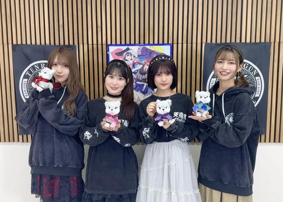 Four young women are posing together, each holding a small plush toy dog with different colored ribbons. They are wearing matching black hoodies with “天狼院” (Tenrouin) printed on them, standing in front of a wooden wall with banners and anime artwork. Their cheerful expressions and coordinated outfits suggest they are part of a promotional event or group.