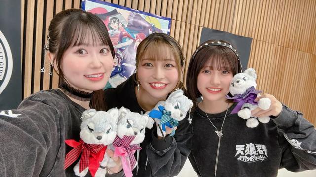 Four young women are posing together, each holding a small plush toy dog with different colored ribbons. They are wearing matching black hoodies with “天狼院” (Tenrouin) printed on them, standing in front of a wooden wall with banners and anime artwork. Their cheerful expressions and coordinated outfits suggest they are part of a promotional event or group.