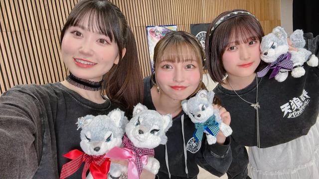 Four young women are posing together, each holding a small plush toy dog with different colored ribbons. They are wearing matching black hoodies with “天狼院” (Tenrouin) printed on them, standing in front of a wooden wall with banners and anime artwork. Their cheerful expressions and coordinated outfits suggest they are part of a promotional event or group.