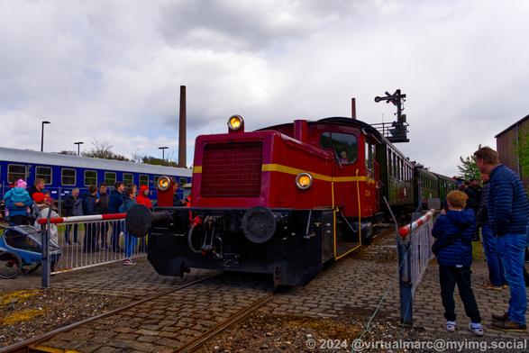 A photo of a red diesel shunting locomotive. carrying passenger wagons, currently driving over the railway crossing.