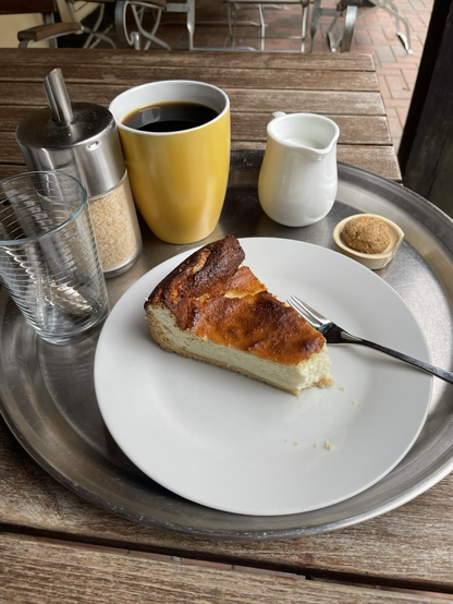 Ein Stück Käsekuchen, dessen Spitze fehlt, liegt auf einem weißen Teller. Am oberen Rand stehen ein Glas, Zucker, ein Pott Kaffee und ein Kännchen Milch.