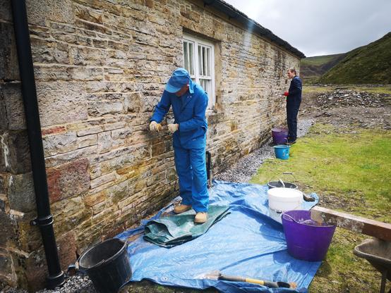 At the rear of a mine workshop, two men are working to repoint the building. One in blue overalls and cap is using a hammer and chisel to rake out old mortar; the second is applying new mortar to the joints between the stones. There is a blue plastic sheet on the ground and buckets with materials and tools lying around.