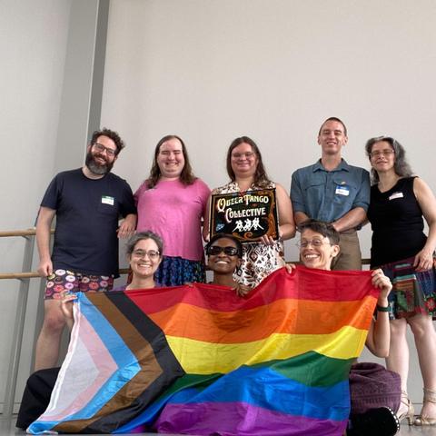8 people, 5 standing in the back row &amp; 3 seated in the front. The center back person holds a “queer tango collective” sign, painted in the fileteado style of Argentina. The front row hold a progress Pride flag.