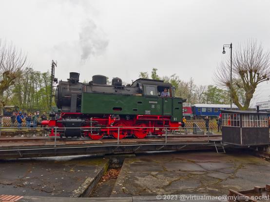 A photo of a dark green steam locomotive from the Hespertalbahn on the turntable. Steam is coming out of the chimney.