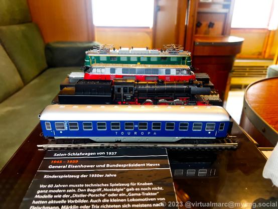 A photo of the inside of a sleeping car from 1937 trhough a window, which was also used by General Eisenhower and Presitend Heuss from 1945 - 1959. There are model railways on the table. From back to front: A green electric train, a red/black V200 diesel locomotive, a steam locomotive and a blue touropa passenger car.