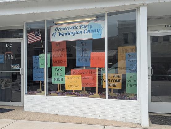 A photograph of a storefront that says Democratic Party of Washington County. There are many protest signs in the window with slogans like "Tax the rich," "We the people have had enough," "all are welcome here," and "we stand with Ukraine."