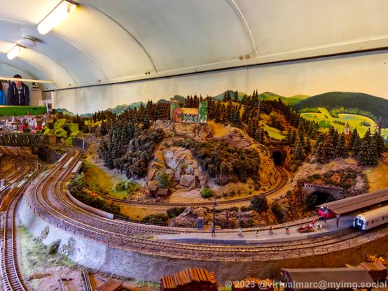 A photo of a model railway with a small train station near some tunnels on a mountainl There is a small castle on the mountain.