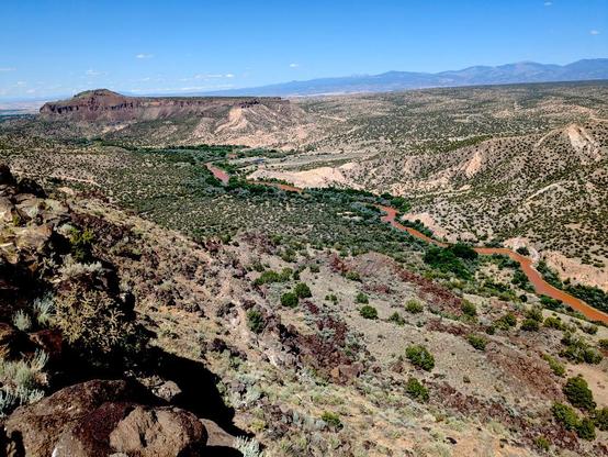 A view of a river winding through a shallow canyon, as seen from the canyon rim. The river is an intense red-brown in color. We're standing on a rocky basalt ledge, and you can see basalt boulders in the slope down to the river; on the other side it's more pale dirt eroded in badlands formations. Both sides of the river have sparse scrubs growing, juniper and sage and similar plants. At the far upstream end of the photo (top left) is a mesa, dark since it's mostly basalt, with a rounded hump on the top.