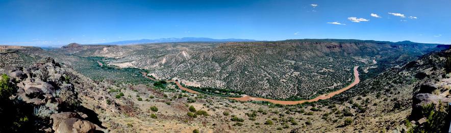 A wide panorama showing a river (the Rio Grande) going through a canyon (White Rock Canyon). We're standing on a cliff looking down at the canyon. To the left, the canyon is shallow and wide, with basalt on this side of the river and badland formations dotted with juniper/sage on the far side. Right of center, the canyon starts to narrow and show steep basalt cliffs on the far side. The water in the river is an intense red-brown color. At the far left of the image, you can see mesas both on this side of the river and on the other side. There's a mountain range in the distance, just left of and above the photo's center. The sky is clear blue with only a few  small puffy clouds.