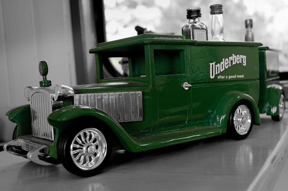 Green vintage-style toy truck with silver accents and shiny wheels, displaying the text "Underberg after a good meal" on its side. In the background, several small glass bottles are visible, placed on a surface against a blurred indoor setting.  
###
Grüner Spielzeugtruck im Vintage-Stil mit silbernen Akzenten und glänzenden Rädern mit dem Schriftzug "Underberg after a good food" an der Seite. Im Hintergrund sind mehrere kleine Glasflaschen zu sehen, die auf einer Fläche vor einem verschwommenen Innenraum platziert sind.