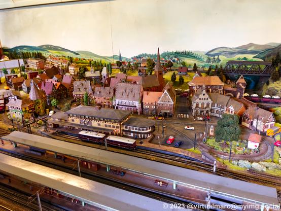 A photo of a model railway. There is a big train station in the front. A city with half-timbered houses and a church is in the back. There is also a tram line in the city.