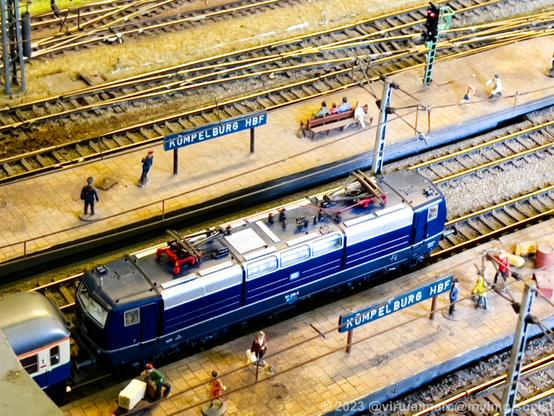 A detail photo of a model railway of a big train station named Kümpelburg Hbf with a blue electric train standing on a platform.