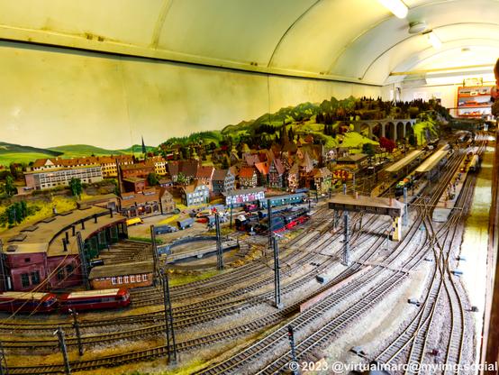 A photo of a model railway with a roundhouse and a railway depot in the front, followed by a big city with half-timbered houses and a big train station. There is a viadukt and a mountain in the back.