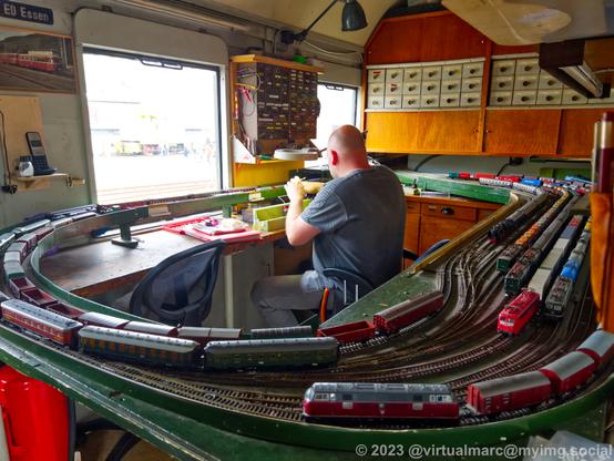 A photo of a back room of a model railway where inactive trains are parked and trains are repaired. A man is sitting in the middle and is repairing a model train.