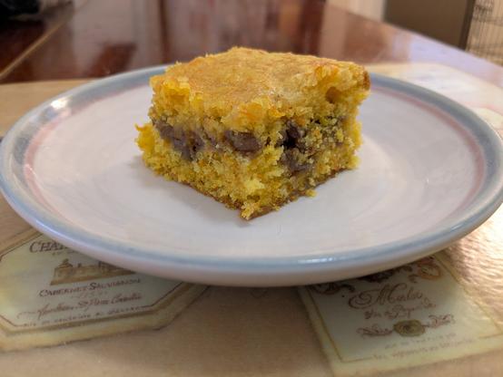 A square piece of cake rests on a small plate. The cake is golden in colour, with a center filling that is a medium brown and contains chopped pecans.