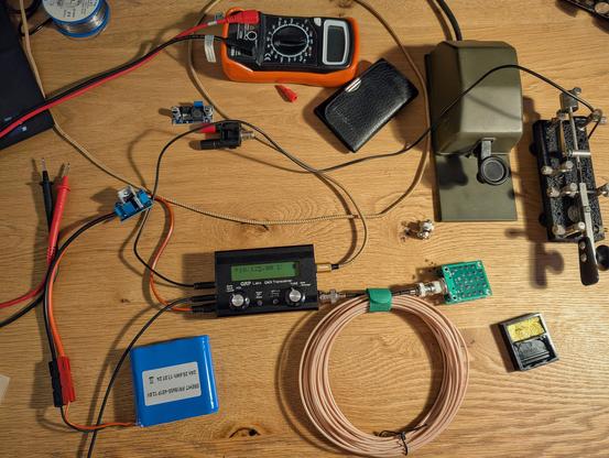 A picture of a desktop with the QMX QRP transceiver sitting in the middle. There are also batteries, a dummy load, morse keys and other tools on it