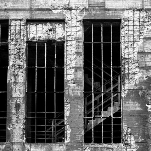 Black and white photo of the outside view of openings in a worn concrete wall with iron frames that formerly held the panes of a latticed window. Behind the windows, a concrete and iron staircase can be seen in the shadow