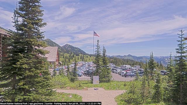 View from the Sunrise Visitor Center in Mount Rainier National Park today. A flagpole holds a flag against a blue sky with drifting clouds. Trees stand in the foreground and along the slopes in the distance. A building appears on the left facing several rows of parked cars. The walkway from the visitor center is in the immediate foreground. It leads to a signpost with information for park visitors.