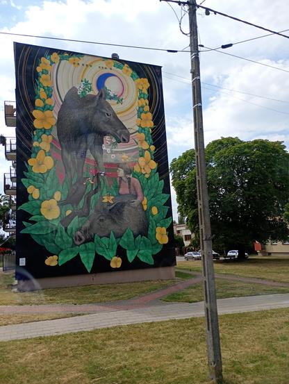 Mural z Simoną Kossak w Białowieży. Na muralu, oprócz Simony, widoczne łoś, dzik i kruk. Wokół zielone liście i żółte kwiaty. Wokół budynku widoczne drzewo, trawa, kostka brukowa, a przed budynkiem słup energetyczny.