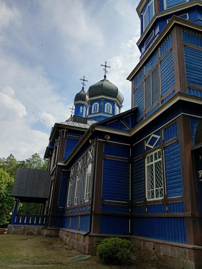 Cerkiew Opieki Matki Bożej w Puchłach elewacja koloru niebieskiego/modrakowego/błękitnego (znawcą nie jestem) z białymi oknami i ciemnym dachem. Wokół widoczne błękitne niebo, trawa i drzewa.