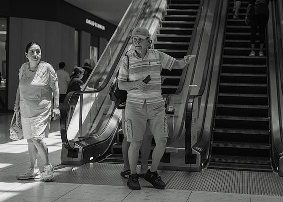 Guy indicating to his wife which way to go, after exiting the escalator at the mall

B&W